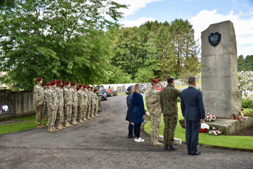 Group of cadets standing while five people pay respects at memorial