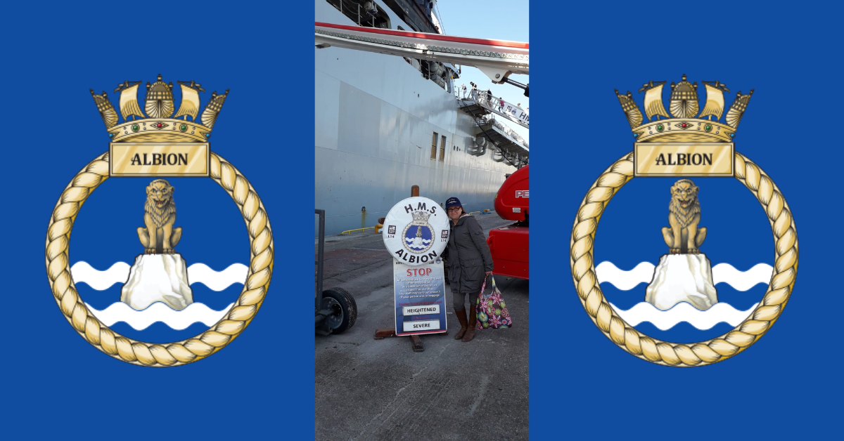 Anne Repo standing next to HMS Albion's sign in front of the vessel with a blue background and two HMS Albion logos on both sides of the image