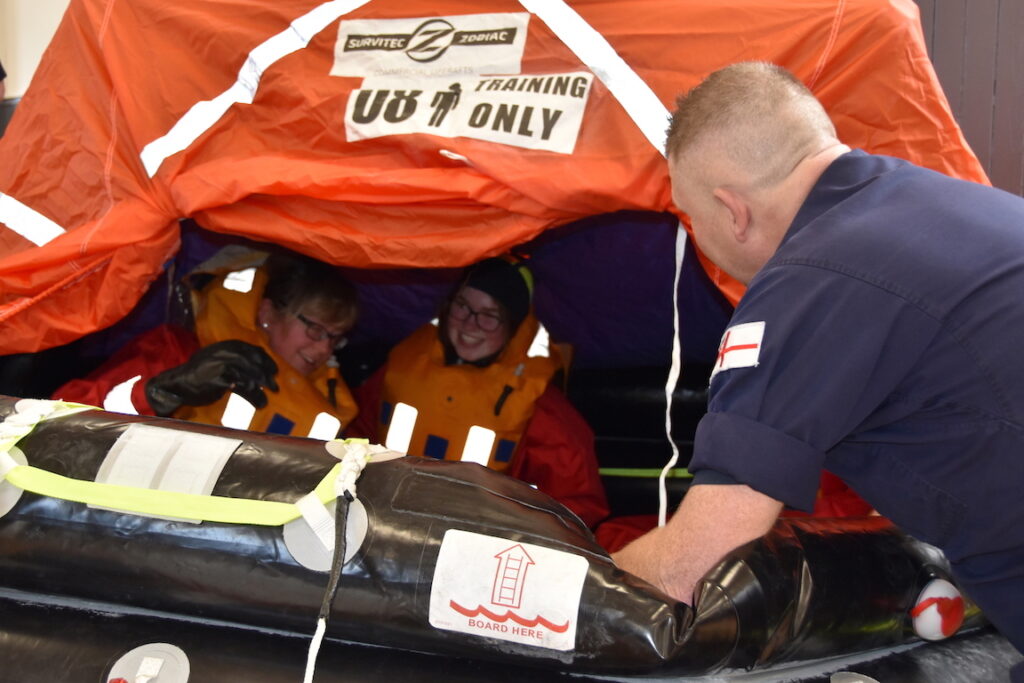 Event participants sit inside a life raft as a sailor looks in.