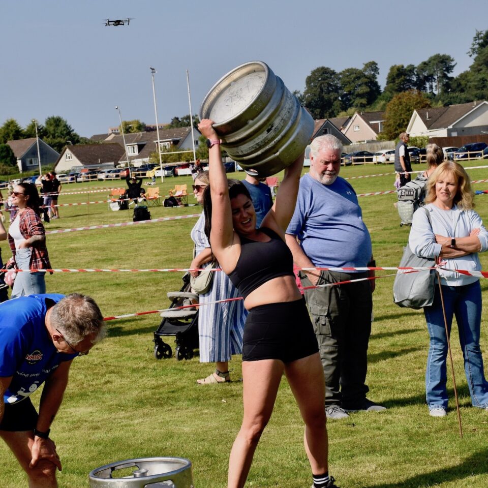 A woman lifts a keg as spectators look on as a drone flies in the background.