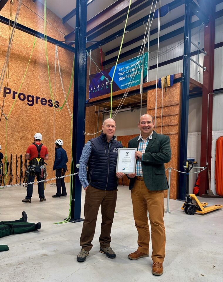Andy Elrod and Ray Watt hold the Armed Forces Covenant certificate with riggers working on a training wall behind.