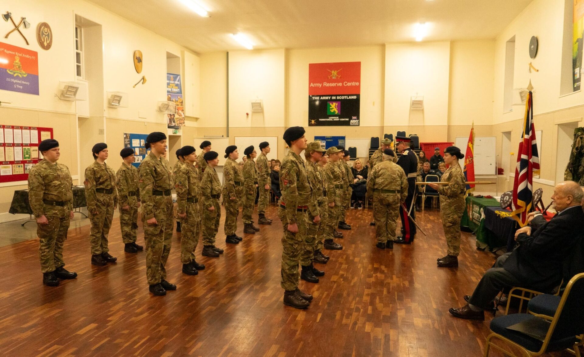 Three rows of Army Cadets in a row in front of two Adult Volunteers, an Army Cadet, the Lord-Lieutenant of Shetland and two seated guests with an audience.