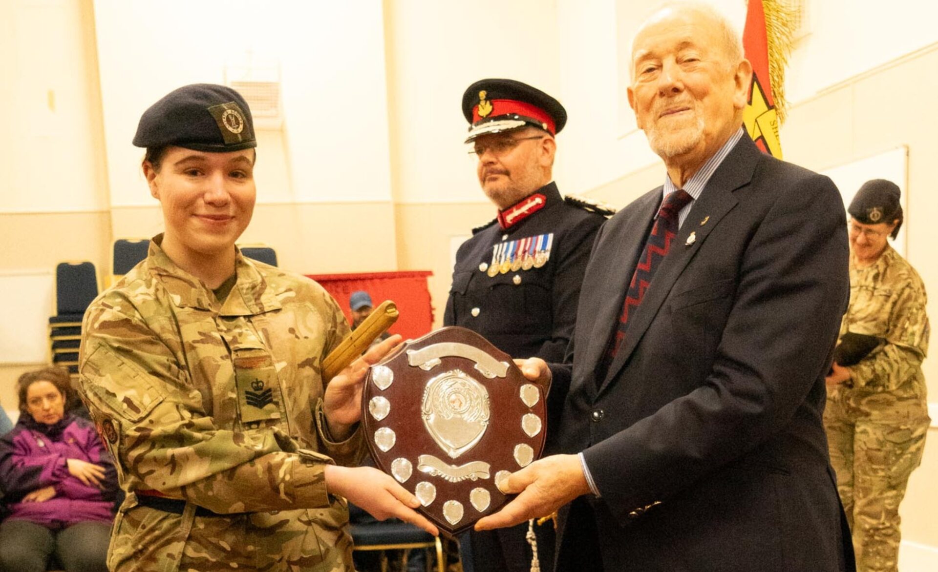 An Army Cadet and a guest holding a shield award in front of the Lord-Lieutenant of Shetland, an Adult Volunteer and a seated audience.